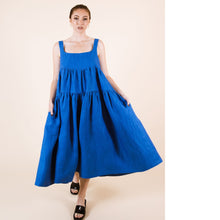 Load image into Gallery viewer, Lady wears a Celestia Dress with square neckline and tiered sections, with hands in pockets, holding the width of the skirt open