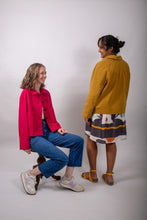 Load image into Gallery viewer, Two models wearing the Jan Jacket, a loose-fitting jacket with sunburst tucks around the neckline, and patch pockets, one worn open, sat down, the other model stands with their back facing the camera