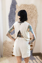 Load image into Gallery viewer, Model's back, wearing a sleeveless top with a central back fastening at the neckline, reveals a cut out of the lower back. Worn with elasticated shirts with wide scoop side pockets.