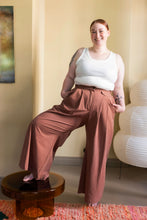 Load image into Gallery viewer, Model with a leg on a stool, wears wide-leg trousers, with pleats at waist and slanted pockets towards the side