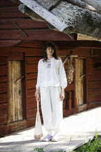 Load image into Gallery viewer, Model walks in front of cabins, wearing a long sleeve top with embroidered centre-front