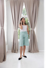 Load image into Gallery viewer, Model stands in front of window wearing knee-length shorts with pleats at waist