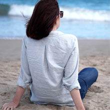 Load image into Gallery viewer, Lady sits on beach wearing Anna shirt, back view shows inverted pleat at yoke.