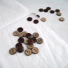 Load image into Gallery viewer, A display of 2-hole coconut buttons with the various natural pattern of two colours, strewn across a piece of fabric