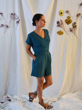 Load image into Gallery viewer, Lady wearing an asymmetrical, 3-buttoned top and shorts playsuit in a green fabric with pleats detail at front shoulder. Lady stood up with hands in pockets.