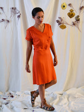Load image into Gallery viewer, Front view of Lady wearing a 3-button asymmetrical top fastening knee length dress in an orange fabric with pleats detail at front shoulder.