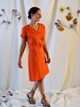 Load image into Gallery viewer, Side view of lady wearing a 3-button asymmetrical top fastening knee length dress in an orange fabric with pleats detail at front shoulder.