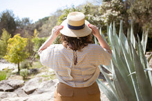 Load image into Gallery viewer, Back view of lady wearing a pleated top with yoke fastened at centre-back with ties, holding onto hat on head
