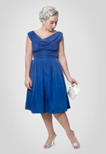 Load image into Gallery viewer, Lady standing wearing sleeveless royal blue dress with cowl neckline, belted waist, pleats from waist, knee length.