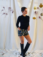 Load image into Gallery viewer, Front view of lady standing, wearing floral pattern curved Pippa shorts, with a plain black top, and knee length black boots.