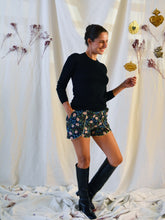 Load image into Gallery viewer, Front view of lady standing, wearing floral pattern curved Pippa shorts, with a plain black top, and knee length black boots.