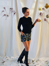 Load image into Gallery viewer, Side view of lady standing, wearing floral pattern curved Pippa shorts, with a plain black top, and knee length black boots.