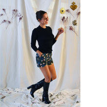 Load image into Gallery viewer, Lady standing wears floral pattern curved Pippa shorts, with a plain black top, and knee length black boots.