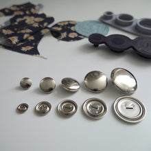 Load image into Gallery viewer, Five different sized metal cover button blanks displayed next to cover tool and circular pieces of fabric