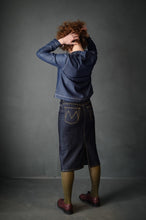Load image into Gallery viewer, Back view of lady wearing the denim below-knee-length pencil skirt. Worn with olive green tights to highlight yellow top stitching of skirt seams, and a denim jacket.