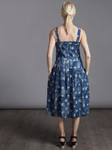 Load image into Gallery viewer, Back view of lady standing, wearing strap top, invisible zipper along centre back seam. Skirt pleated in at waistline, dress length below the knee. Dress in a blue and white round pattern fabric. Lady has hands in pockets.