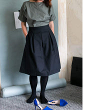 Load image into Gallery viewer, Lady wears a black three pleat skirt. Worn with black tights, blue heels kicked off.