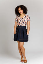Load image into Gallery viewer, Lady stands wearing a denim, short Brumby Skirt with contrasting topstitching detail. Hands in deep scoop pockets.