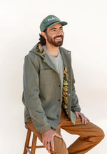 Load image into Gallery viewer, Model sat on stool wears a hooded Beachcomber Jacket with elasticated cuffs, and patterned lining
