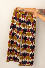 Load image into Gallery viewer, Hands holding up a pair of Flâneur trousers, back view, in busy print fabric, with patch pockets, and elasticated waist
