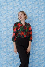 Load image into Gallery viewer, Model wearing a colourful floral blouse made with La Roseraie Viscose fabric, stood against a patterned background
