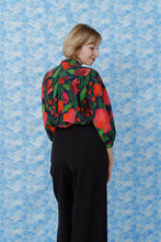 Load image into Gallery viewer, Back view of model wearing a colourful floral blouse, made with La Roseraie Viscose fabric, stood in front of a patterned background
