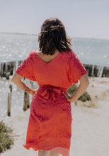 Load image into Gallery viewer, Back view of lady at the beach with hands on hips wears the Transat Dress: a V-neck back, short sleeved, knee length dress with waist bow tie at back
