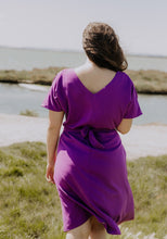 Load image into Gallery viewer, Back view of lady at the beach wearing the Transat Dress: a V-neck, short sleeved, knee length dress with waist tie
