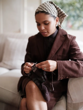 Load image into Gallery viewer, Model sits on couch knitting in a cord Blazer and patterned headscarf
