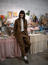 Load image into Gallery viewer, Model in a brown suit standing in a room with tables and various items.
