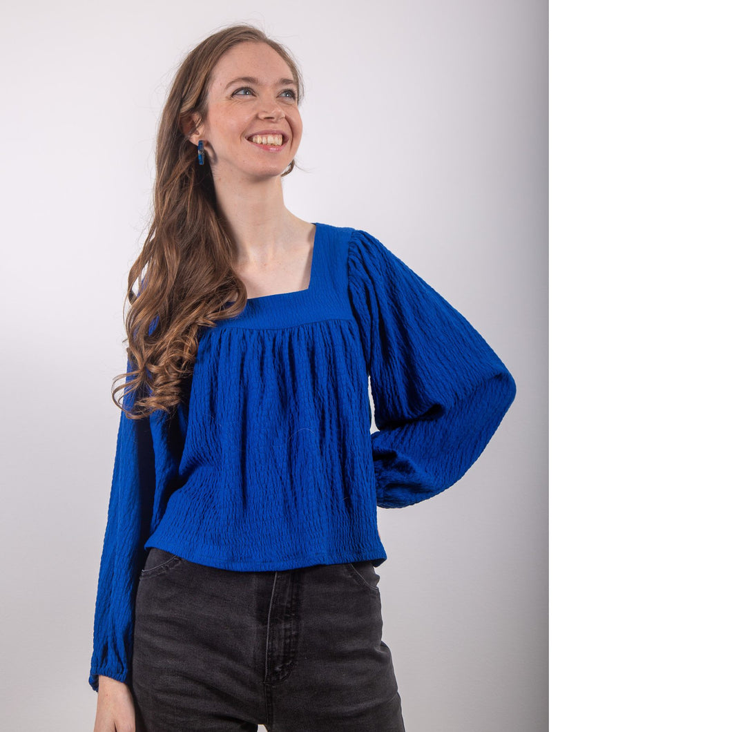 Model stands wearing a textured blouse with a square neckline and voluminous long sleeves