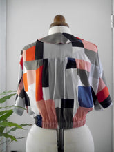 Load image into Gallery viewer, A short-sleeve collared shirt with a colourful geometric patchwork print, shown from the back on a mannequin.
