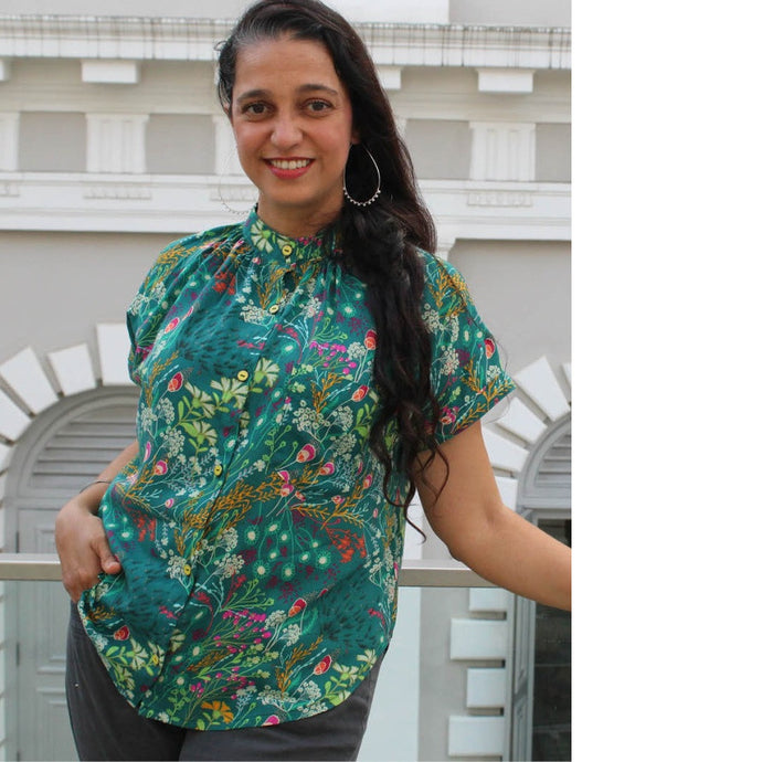 Lady leans on wall, wears a Sille Shirt made in floral print fabric, shoes gathers at collar stand neckline, and button front.