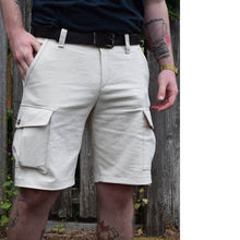 Load image into Gallery viewer, Man wears cream coloured cargo shorts with the patch pockets with flaps on the sides. Worn with a leather belt.
