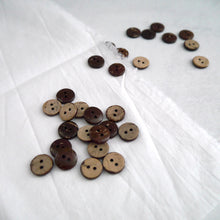 Load image into Gallery viewer, A display of 2-hole coconut buttons with the various natural pattern of two colours, strewn across a piece of fabric
