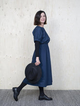 Load image into Gallery viewer, Side view of lady wearing denim blue mid-calf length dress, elasticated at cuffs and at waist, holding a hat.
