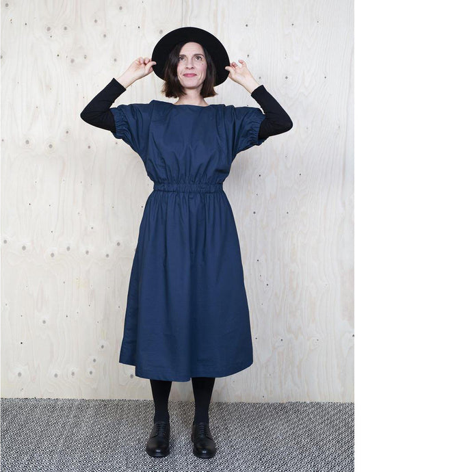 Lady wearing dress with elasticated cuff and waist detail in a denim like fabric, layered over a black jersey top and black tights.