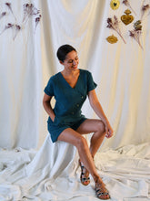 Load image into Gallery viewer, Lady wearing an asymmetrical, 3-buttoned top and shorts playsuit in a green fabric with pleats detail at front shoulder, laughing.
