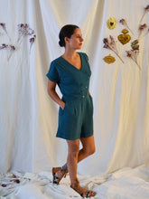 Load image into Gallery viewer, Lady wearing an asymmetrical, 3-buttoned top and shorts playsuit in a green fabric with pleats detail at front shoulder. Lady stood up with hands in pockets.
