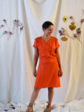 Load image into Gallery viewer, Lady wearing a 3-button asymmetrical top fastening knee length dress in an orange fabric with pleats detail at front shoulder.
