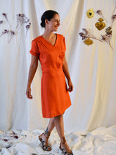 Load image into Gallery viewer, Side view of lady wearing a 3-button asymmetrical top fastening knee length dress in an orange fabric with pleats detail at front shoulder.
