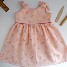 Load image into Gallery viewer, A baby dress, with knot-tie shoulders, and gathered at waist, made with Organic Cotton Double Gauze Melons Fabric, displayed on table.

