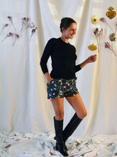 Load image into Gallery viewer, Front view of lady standing, wearing floral pattern curved Pippa shorts, with a plain black top, and knee length black boots.
