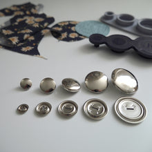 Load image into Gallery viewer, Five different sized metal cover button blanks displayed next to cover tool and circular pieces of fabric
