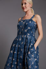 Load image into Gallery viewer, Angled view of lady standing, wearing strap top with four buttons down centre front, pleated in skirt at waistline, dress length below the knee. Dress in a blue and white round pattern fabric. Lady has hands in pockets.
