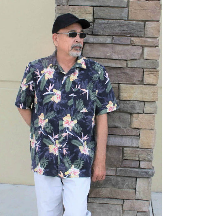 Man stands in front of wall wearing a Tropical print short sleeve shirt, with one chest pocket. Worn with light coloured jeans, black baseball cap and sunglasses.