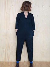 Load image into Gallery viewer, Lady wears a V-neck jumpsuit in blue fabric. Three quarter length sleeves, and tapered legs. Lady stands in front of wall with head turned to the left.

