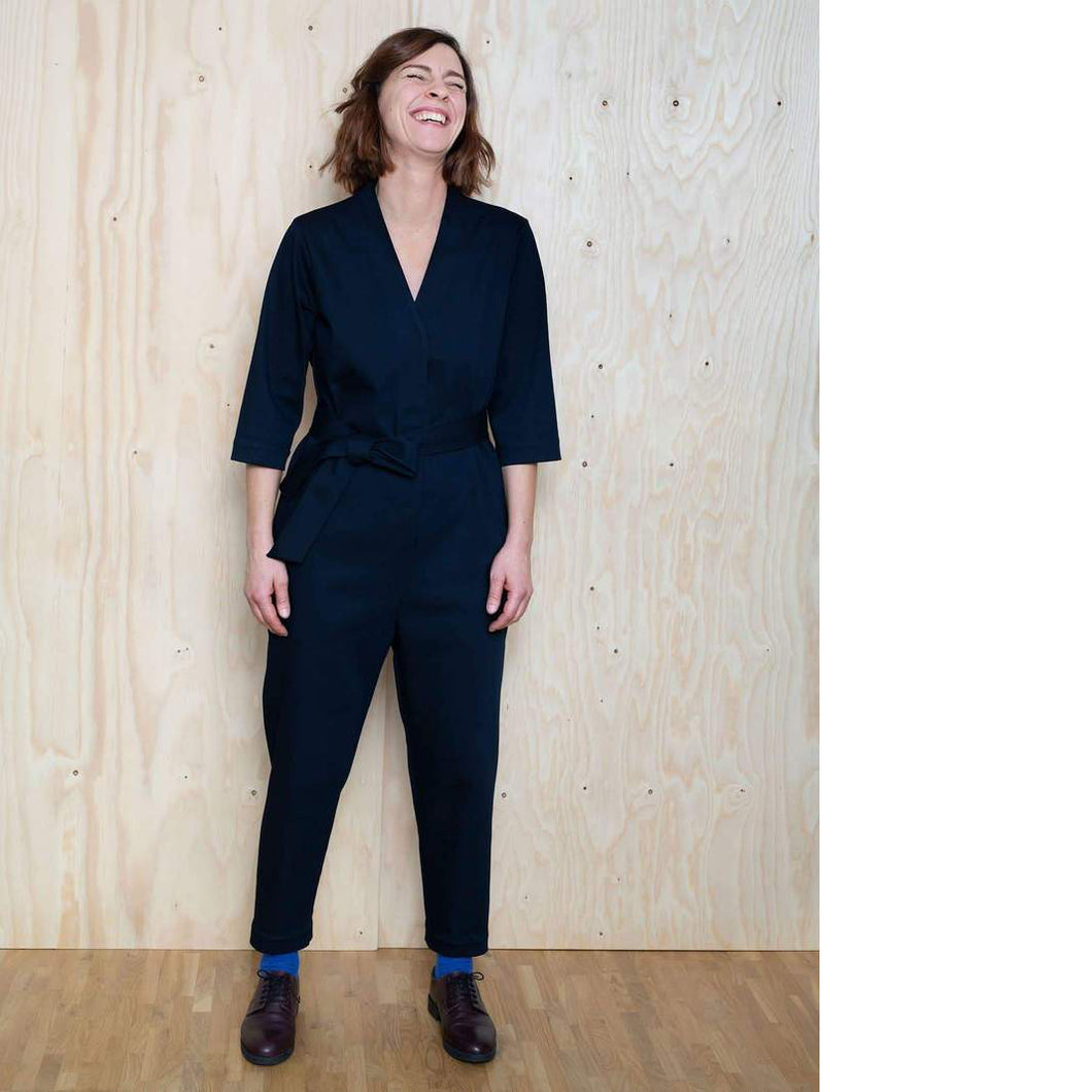 Lady stands in front of wall laughing. Lady wears a V-Neck jumpsuit with tie at waist, narrow fit trouser.
