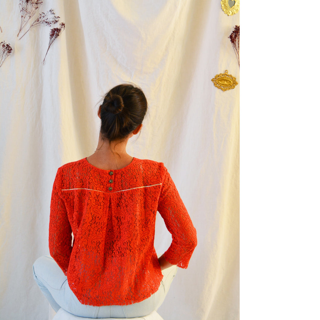 Back view of lady sat down cross legged, wearing an orange lace long sleeve top with three button fastening at neckline, which holds an inverted pleat down along the centre back.