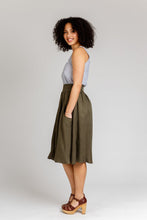 Load image into Gallery viewer, Lady stands wearing a gathered, knee-length Brumby skirt with hands in deep scoop pocket, side view.
